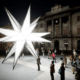 La plaza Sant Jaume se convierte este año en un verdadero epicentro de la Navidad en Barcelona, gracias a una propuesta innovadora que combina tradición e innovación. La instalación de una espectacular estrella luminosa de 20 puntas, llamada "Origen", será el centro de atención de la plaza, mientras que dos pesebres, situados en el interior del Ayuntamiento y del Palau de la Generalitat, complementarán la decoración navideña. Este proyecto, fruto de la colaboración entre el Ayuntamiento de Barcelona y la Generalitat de Catalunya, promete ofrecer una experiencia única para ciudadanos y visitantes.

Una estrella monumental como símbolo de perfección
La estrella "Origen" no es solo una pieza decorativa; es una obra de arte que destaca por su diseño y significado. Creada por Xevi Bayona y Àlex Posada, la estrella representa un icosaedro, una figura geométrica de 20 caras triangulares que simboliza perfección, armonía y equilibrio. Este diseño geométrico también evoca conceptos universales, como el momento germinal del universo, conocido como el Big Bang, cuando la oscuridad se transformó en luz y energía.

Con un diámetro de nueve metros y un peso de 2,5 toneladas, la estrella está fabricada con hierro, metacrilato y tecnología LED, lo que garantiza tanto su durabilidad como su espectacularidad visual. Durante el día, esta obra se presenta como una imponente escultura geométrica en el lateral de la plaza, cerca de las calles Ferran y del Call. Sin embargo, es al anochecer cuando despliega todo su potencial: se convierte en un faro luminoso que proyecta reflejos de luz sobre las fachadas del Ayuntamiento y del Palau de la Generalitat.

Además, la instalación cuenta con una banda sonora original que añade un componente sensorial al espectáculo, haciendo que la experiencia sea aún más inmersiva y cautivadora.

Dos pesebres para honrar la tradición navideña
La decoración de la plaza Sant Jaume no estaría completa sin los tradicionales pesebres, que este año se han trasladado al interior de los edificios históricos para enriquecer la experiencia de los visitantes. El Pati de Carruatges del Ayuntamiento acogerá el pesebre tradicional, elaborado por la Associació de Pessebristes. Este diorama incluirá referencias al centenario del poeta Joan Salvat-Papasseit, quien trabajó en el Moll de la Fusta, y a la reciente edición de la Copa América de vela en Barcelona. La exposición estará abierta al público del 29 de noviembre al 5 de enero, ofreciendo una oportunidad única para disfrutar de esta emblemática tradición en un entorno singular.

Por su parte, el Palau de la Generalitat abrirá sus puertas entre el 14 y el 20 de diciembre para mostrar su decoración navideña. Los visitantes podrán descubrir un pesebre, un tió y avetos decorados, ambientados con un estilo que combina tradición y modernidad. Esta iniciativa no solo fomenta el espíritu navideño, sino que también permite a la ciudadanía explorar espacios habitualmente reservados.

Colaboración entre instituciones para una Navidad especial
El proyecto de decoración de la plaza Sant Jaume es el resultado de una estrecha colaboración entre el Ayuntamiento y la Generalitat, que han trabajado juntos para ofrecer una propuesta unificada y coherente. Ambas instituciones también han compartido la misma ornamentación vegetal en los balcones de sus edificios, reforzando la idea de cohesión.

El coste total de la estrella luminosa asciende a 145.000 euros, mientras que el pesebre del Ayuntamiento ha supuesto una inversión adicional de 50.000 euros. Aunque el Ayuntamiento ha asumido la mayor parte del gasto, el concejal Jordi Marcé destacó la importancia de esta colaboración: "Estamos agradecidos de que la Generalitat nos haya apoyado en una iniciativa que siempre ha sido del Ayuntamiento. Esto refuerza el compromiso de ambas instituciones con la Navidad y con la ciudad de Barcelona."

Una instalación con visión de futuro
La estrella "Origen" no está concebida como una decoración efímera. Tanto el Ayuntamiento como la Generalitat han dejado abierta la posibilidad de reutilizar esta monumental pieza en futuras ediciones, consolidándola como un símbolo navideño de Barcelona. Este enfoque no solo busca maximizar el impacto de la inversión, sino también establecer un vínculo emocional con los ciudadanos, que podrán disfrutar de esta obra año tras año.

Polémica y tradición en Sant Jaume
La decoración navideña de la plaza Sant Jaume siempre ha sido objeto de debate, especialmente en torno a los pesebres. El traslado del pesebre al interior del Ayuntamiento y la Generalitat ha generado opiniones divididas, pero las instituciones han defendido la decisión como una forma de enriquecer la experiencia navideña. Según Marcé, "reubicamos las piezas para aprovechar al máximo el espacio y ofrecer una propuesta artística más completa."

La estrella luminosa y los dos pesebres no solo buscan embellecer la plaza, sino también reforzar el mensaje de unidad y colaboración entre las instituciones. Además, el diseño y la ubicación de la estrella fueron seleccionados para resaltar el carácter icónico de Sant Jaume como punto de encuentro entre tradición e innovación.

Sant Jaume, el corazón de la Navidad en Barcelona
Con estas iniciativas, la plaza Sant Jaume se consolida como uno de los principales puntos de interés navideño en Barcelona. La combinación de una monumental estrella luminosa, dos pesebres emblemáticos y una decoración coherente en toda la plaza crea un entorno mágico que invita a barceloneses y turistas a sumergirse en el espíritu de las fiestas.

Sant Jaume no solo es un espacio de celebración, sino también un símbolo de la capacidad de Barcelona para combinar tradición y modernidad, demostrando que la Navidad puede ser un momento para innovar sin perder de vista las raíces culturales de la ciudad.